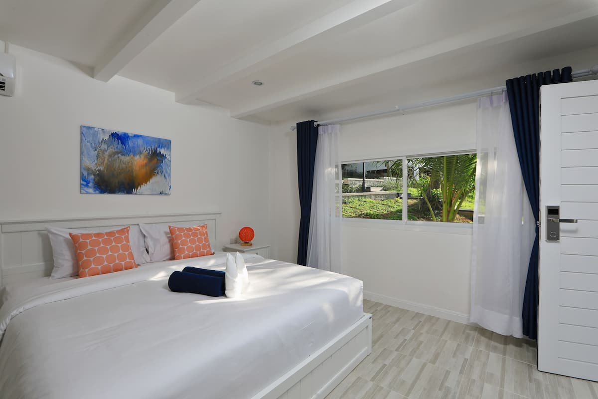 A bedroom with a large bed, white walls, a painting, a window with curtains, and a view of greenery outside.