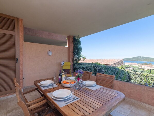 Mesa de madera dispuesta para dos personas, con platos, cubiertos y un frutero en un balcón con vistas al mar.