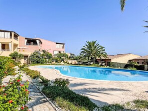Una piscina rodeada por un jardín con diversas plantas y una palmera. Se observan edificios con balcones al fondo.