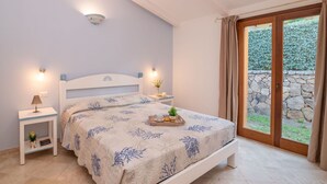 Un dormitorio con cama, mesitas de noche, una pequeña planta y una bandeja con aperitivos. Se ve una ventana con cortinas y vistas al verdor exterior.