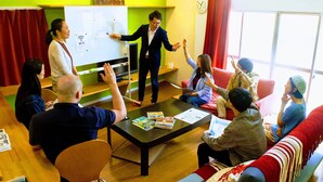 A group of people in a meeting room with a whiteboard, a table with magazines, and a red couch. - Guesthouse LuLuLu Atago (Kochi)