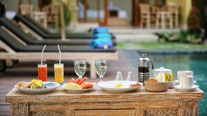 Ein Frühstückstisch mit verschiedenen Speisen und Getränken, einer Kaffeekanne und im Hintergrund ein Blick auf einen Pool und Liegestühle.