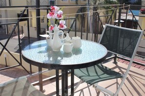 A round glass table with a white pitcher, teapot, and cups on a balcony with metal railings and a chair.