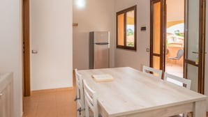 Une cuisine compacte dotée d’un réfrigérateur intégré, d’une table à manger avec des chaises, et offrant une vue sur une terrasse extérieure par une porte ouverte.