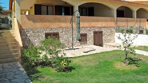 Une maison à deux étages avec une façade en pierre, un patio couvert et un petit jardin agrémenté d’arbustes et d’un arbre.