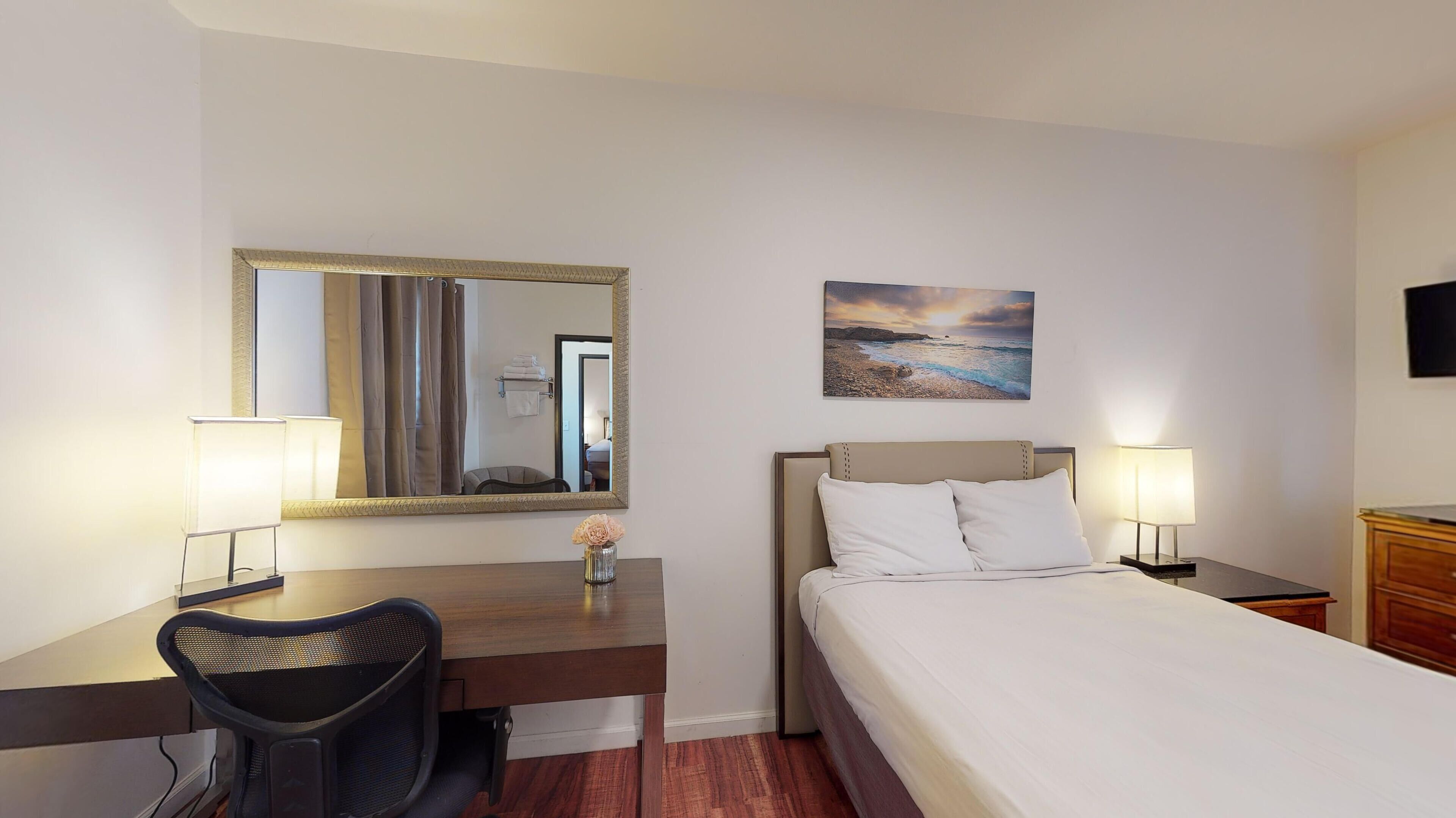 Une chambre d’hôtel avec un lit, un bureau et une chaise, un miroir, un tableau et un téléviseur fixé au mur.
