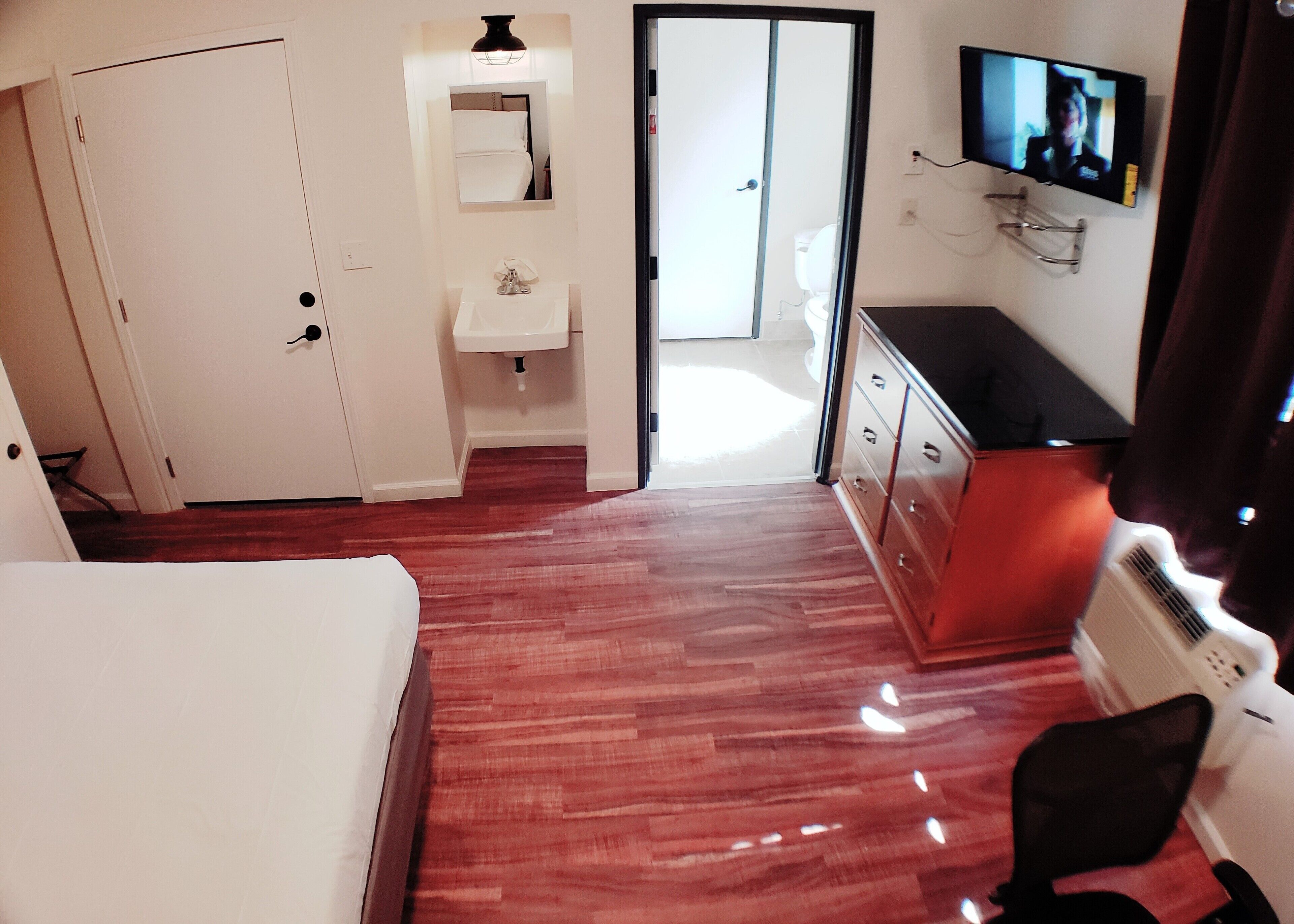 A hotel room with a bed, a desk, a chair, a TV, a mirror, and a bathroom visible through an open door.
