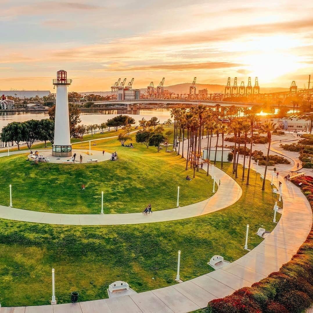 A park with a central monument, pathways, and a waterfront with industrial structures in the background.