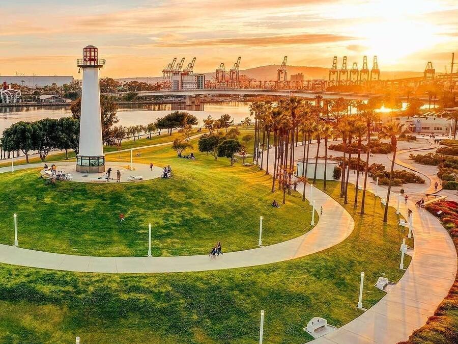 A park with a central monument, pathways, and a waterfront with industrial structures in the background.