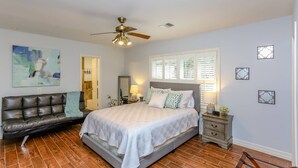 Une chambre à coucher comprenant un lit, une table de chevet, une commode, un canapé en cuir, un ventilateur de plafond et une fenêtre avec des stores.