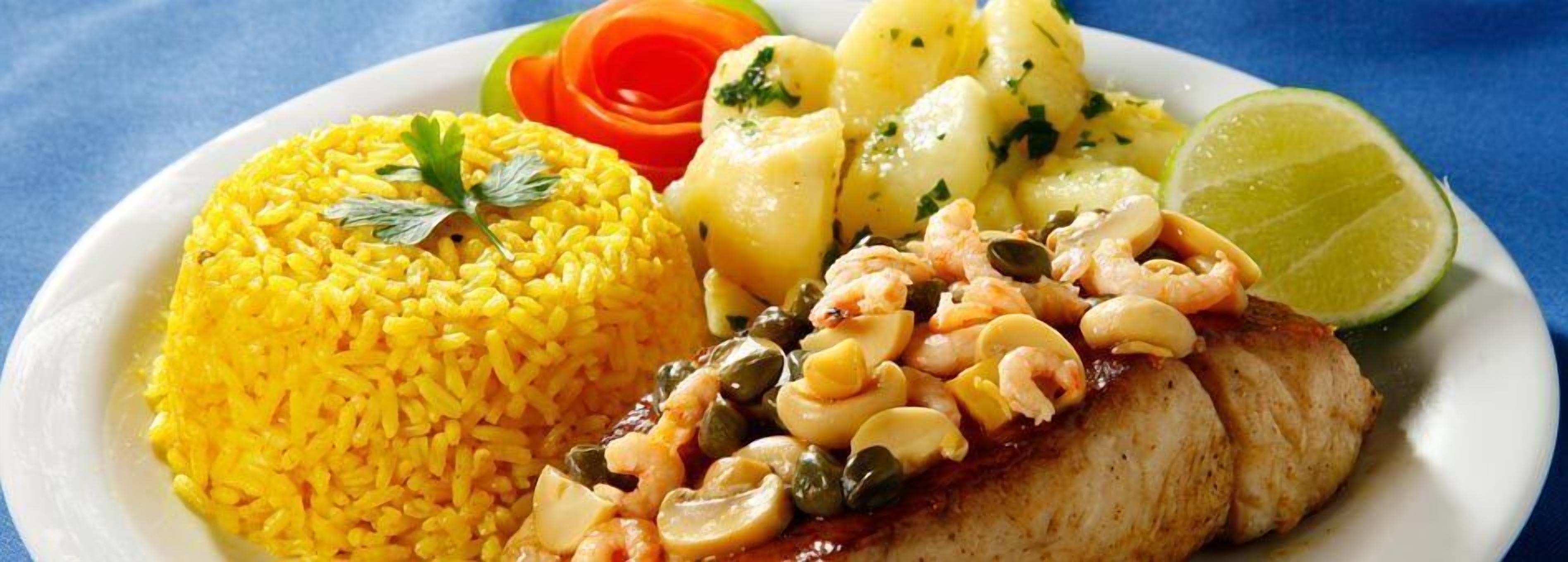 Ein Teller mit gelbem Reis, ein gegrillter Fisch mit Garnelen und Oliven, dazu gekochte Kartoffeln und eine Limettenspalte.