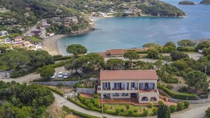 Una villa costera con techo de teja terracota, rodeada de exuberante vegetación y una playa.