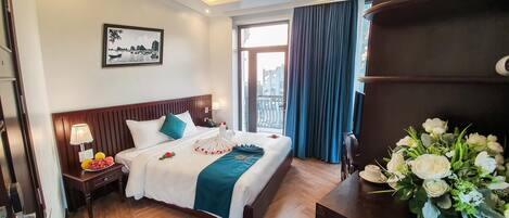 A hotel room with a large bed, a nightstand with a lamp and fruit bowl, a view of the city through a window, and a bouquet of flowers on a table.