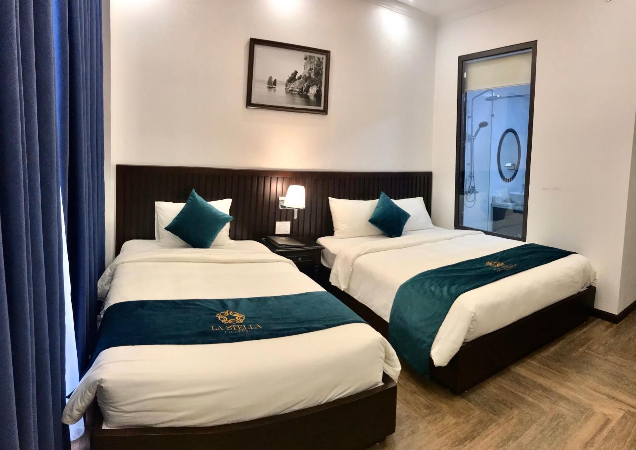 A hotel room with two beds, each with a matching headboard and pillow. A framed picture hangs on the wall above the beds, and a window with a view of the sea is visible.