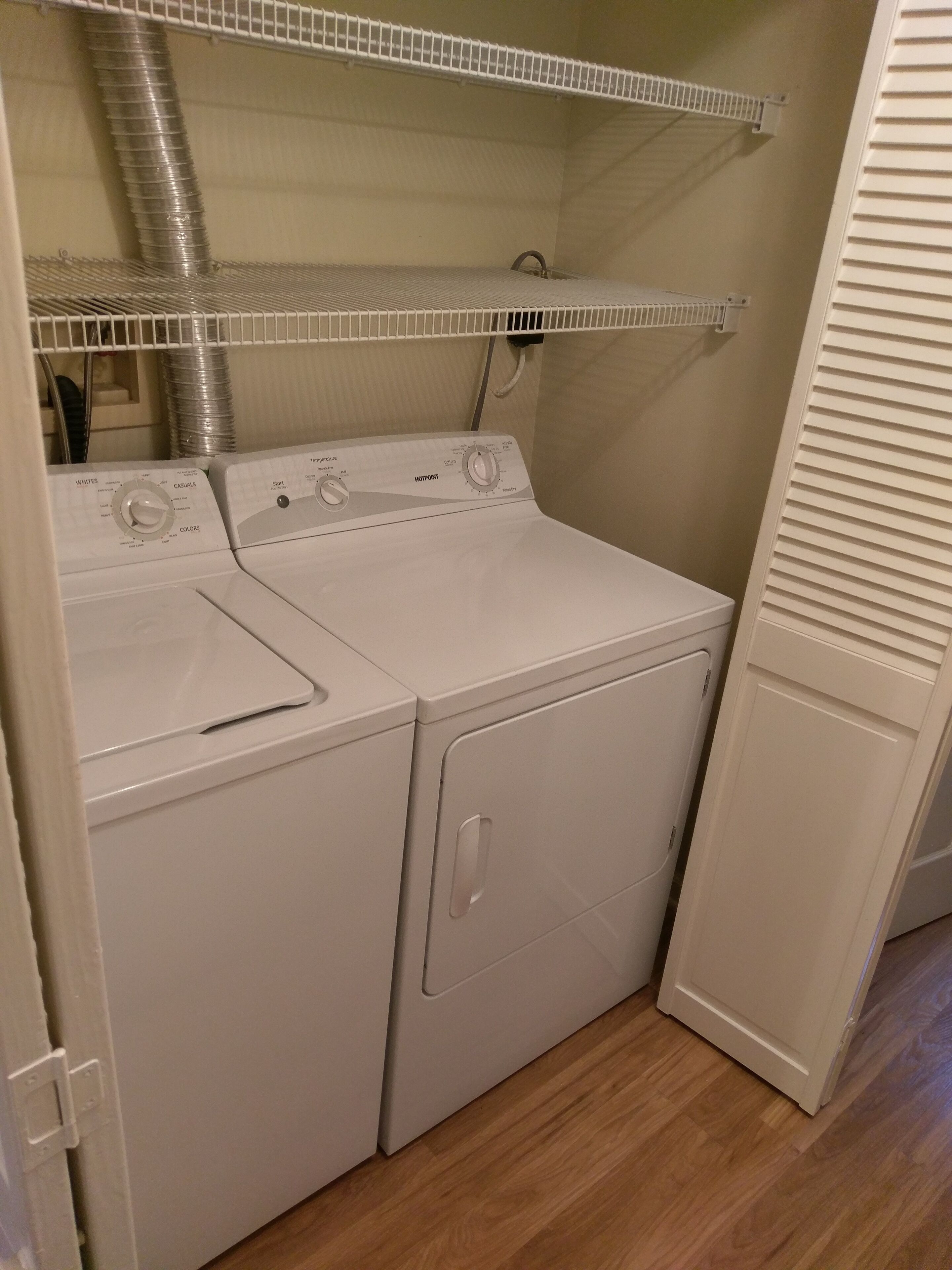 A compact laundry room with a front-loading washer and dryer, a metal vent, and a white door.