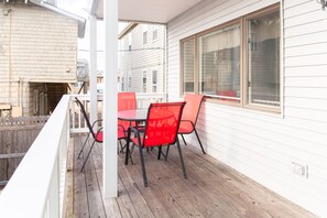 Een overdekte veranda met een houten tafel en rode stoelen, wit beklede gevel en een raam met zonwering.