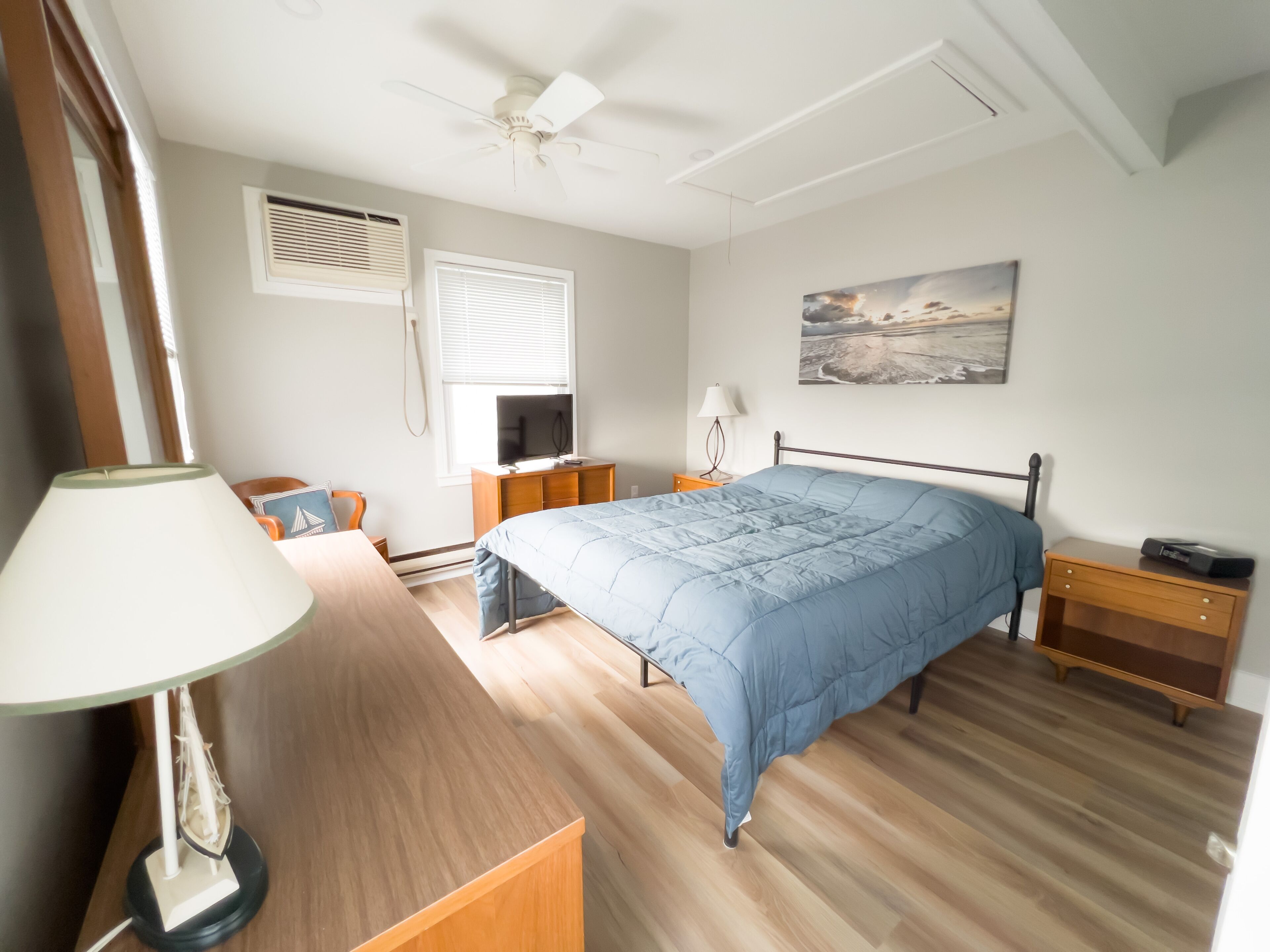 Una camera da letto con un letto, una scrivania, una sedia, una lampada, una TV, un comodino e una finestra con persiane, oltre a una ventola a soffitto.