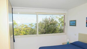 A bedroom with a bed, a window with a view of trees and the sea, and a small bedside table with a lamp. - Nice apartment for 4 people with WIFI, TV and terrace (Portoferraio)
