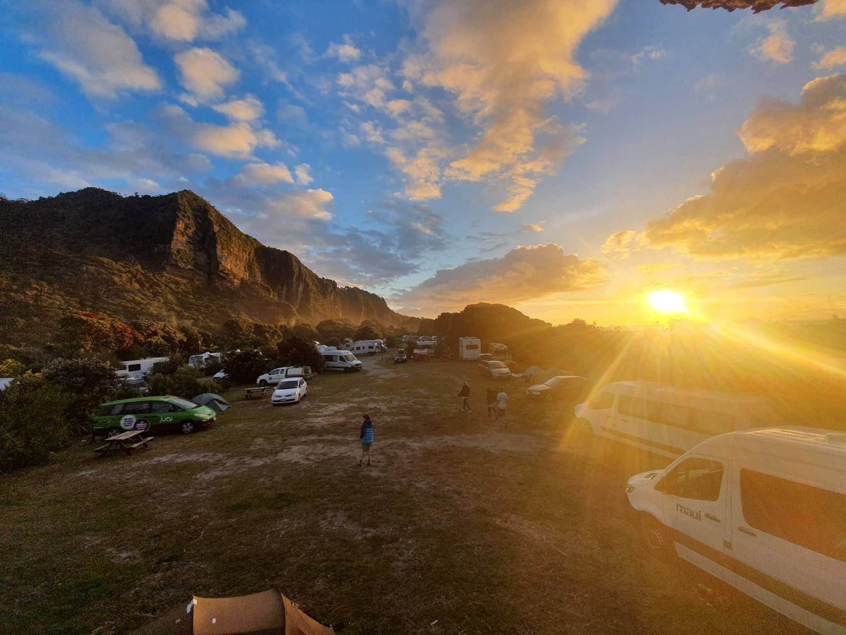 Un camping con vehículos recreativos y automóviles estacionados en un área verde, con montañas al fondo.