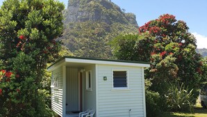 Une petite cabane à un seul étage, dotée d’un porche et d’une plaque portant le numéro « 4 », entourée d’une végétation luxuriante et avec une montagne en arrière-plan.