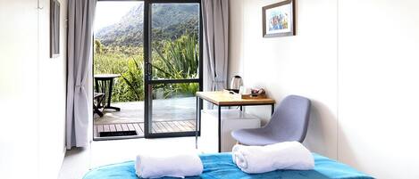 A hotel room with a bed, a chair, a table, and a view of greenery and mountains through a sliding door.