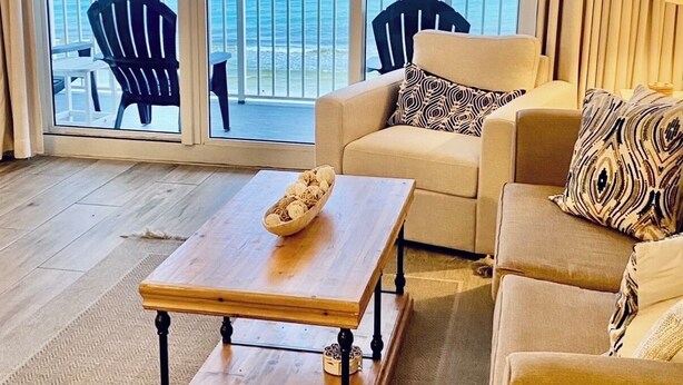 Una sala de estar con una mesa de centro de madera, sofás color beige y vista al océano a través de ventanas amplias.