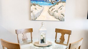 Una mesa de comedor con superficie de vidrio rodeada por sillas de madera con asientos acolchados, un adorno blanco sobre la mesa y un cuadro de una escena de playa en la parte superior.