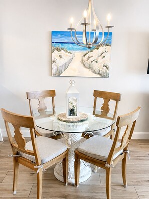 A dining table with a glass top surrounded by wooden chairs with cushioned seats, a white decorative item on the table, and a painting of a beach scene above.