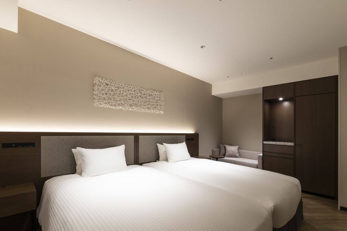 A modern hotel room with a large bed, a bedside table, a nightstand, a seating area with a sofa, and a wardrobe.