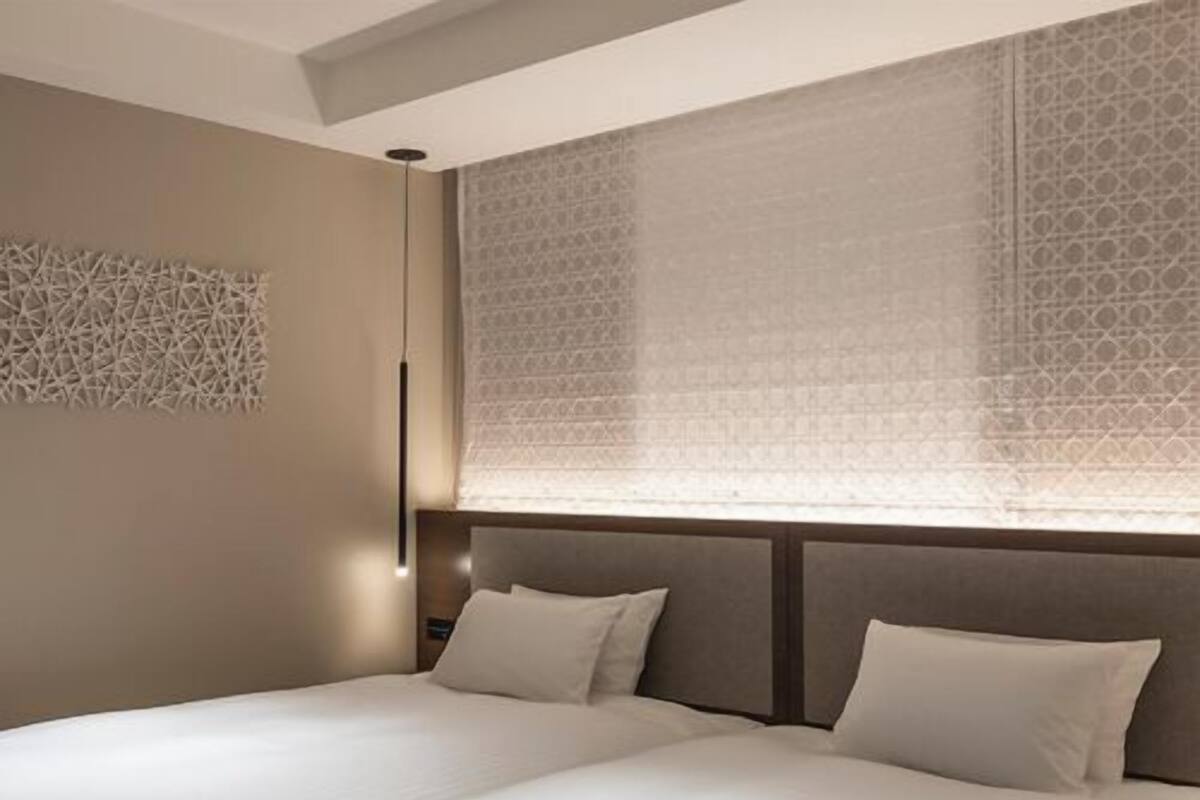 A double bed with a headboard, a wall-mounted light fixture, and a patterned window blind.