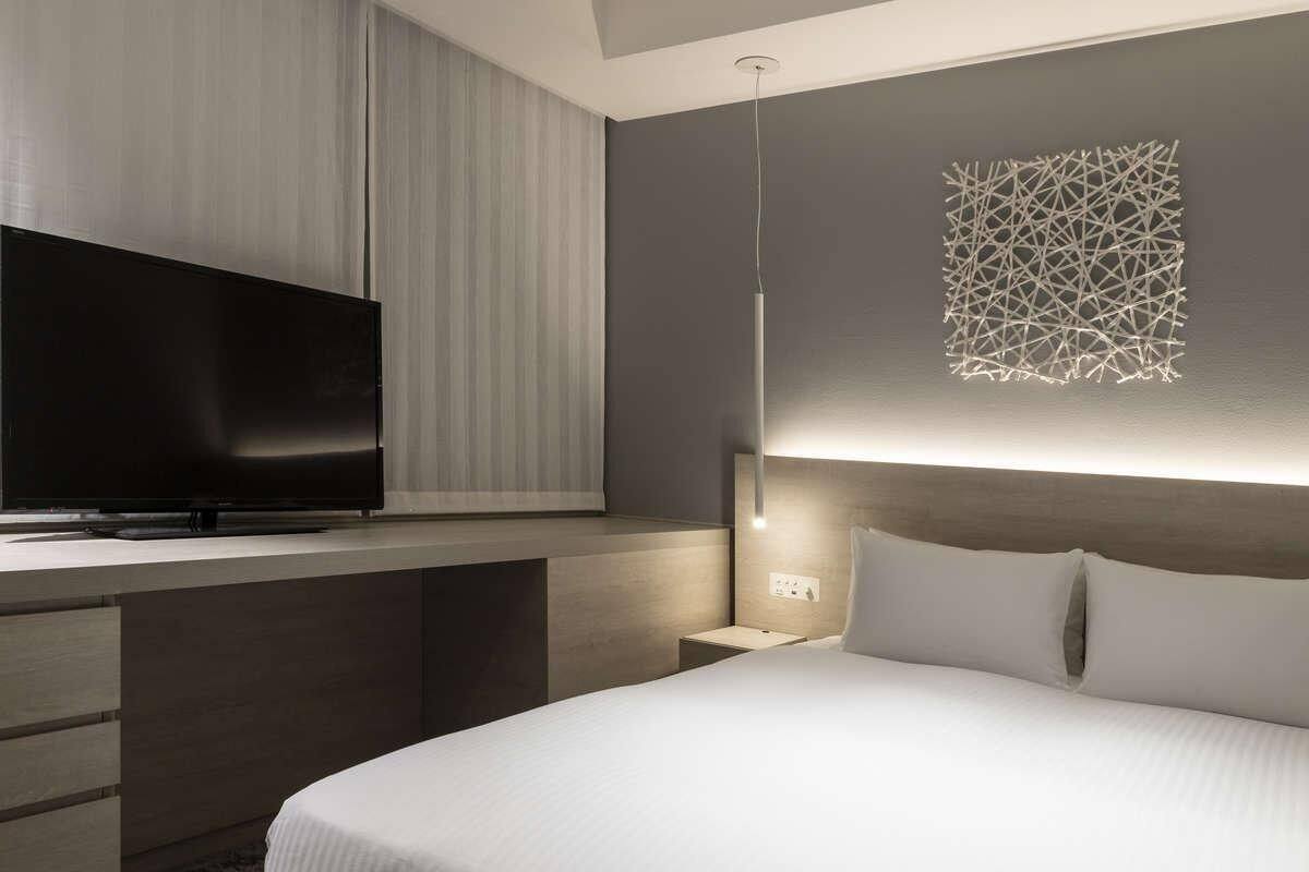 A modern hotel room with a flat-screen TV, a bedside table with a lamp, and a decorative wall piece.