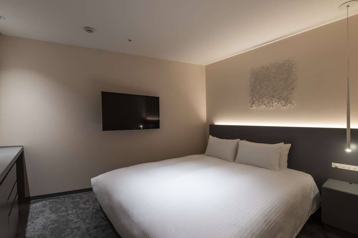 A modern hotel room with a large bed, a flat-screen TV mounted on the wall, and a bedside table with a lamp.