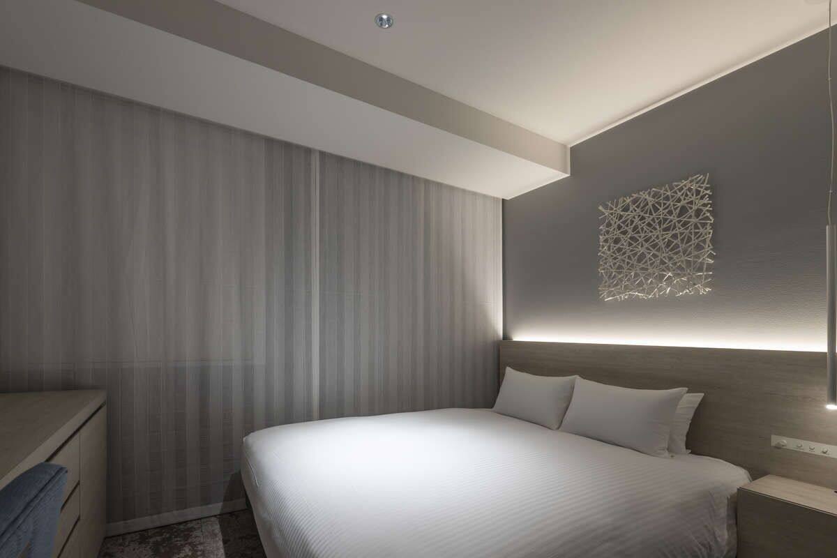 A modern hotel room with a large bed, a nightstand, a dresser, and a wall-mounted light fixture.