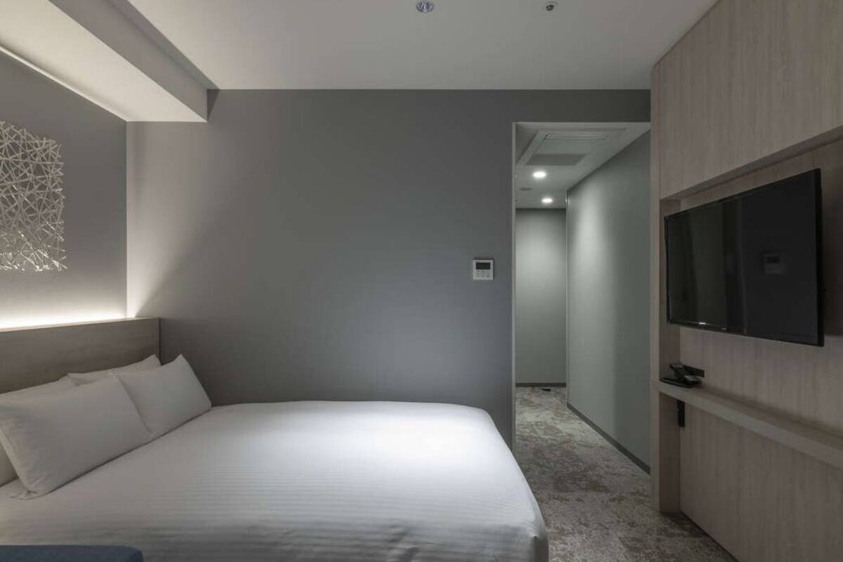 A modern hotel room with a large bed, a flat-screen TV mounted on the wall, and a hallway leading to other rooms.