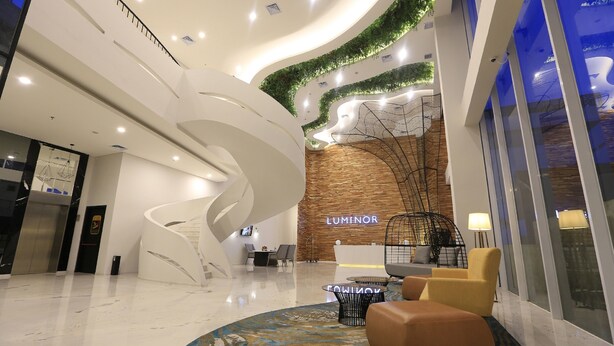 Un hall d’entrée moderne avec un escalier à la forme arrondie, un comptoir d’accueil portant l’inscription « LUMINOR » et un espace salon agrémenté d’un tapis à motifs.