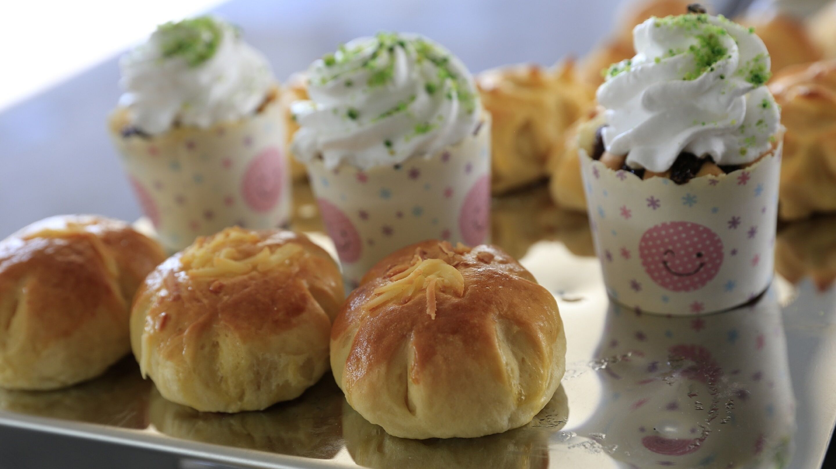 A tray of pastries, including cupcakes with whipped cream and a smiley face design, and small buns.