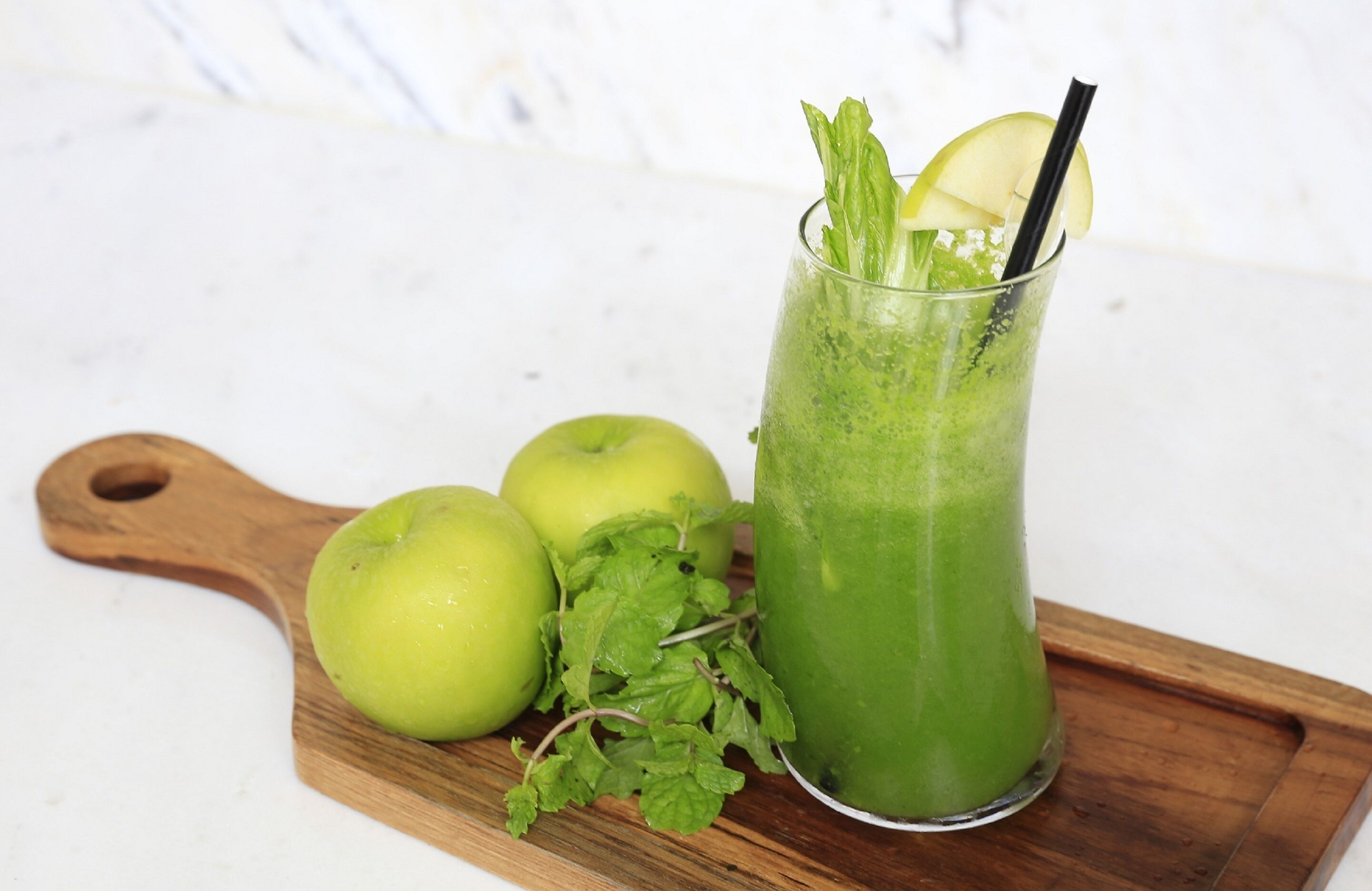 Ein grünes Getränk im Glas mit Strohhalm, begleitet von drei grünen Äpfeln und Minzblättern auf einer Holzplatte.