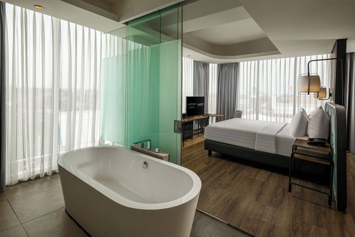Habitación de hotel moderna con una bañera grande, una cama, un televisor y vistas a la ciudad a través de amplios ventanales.