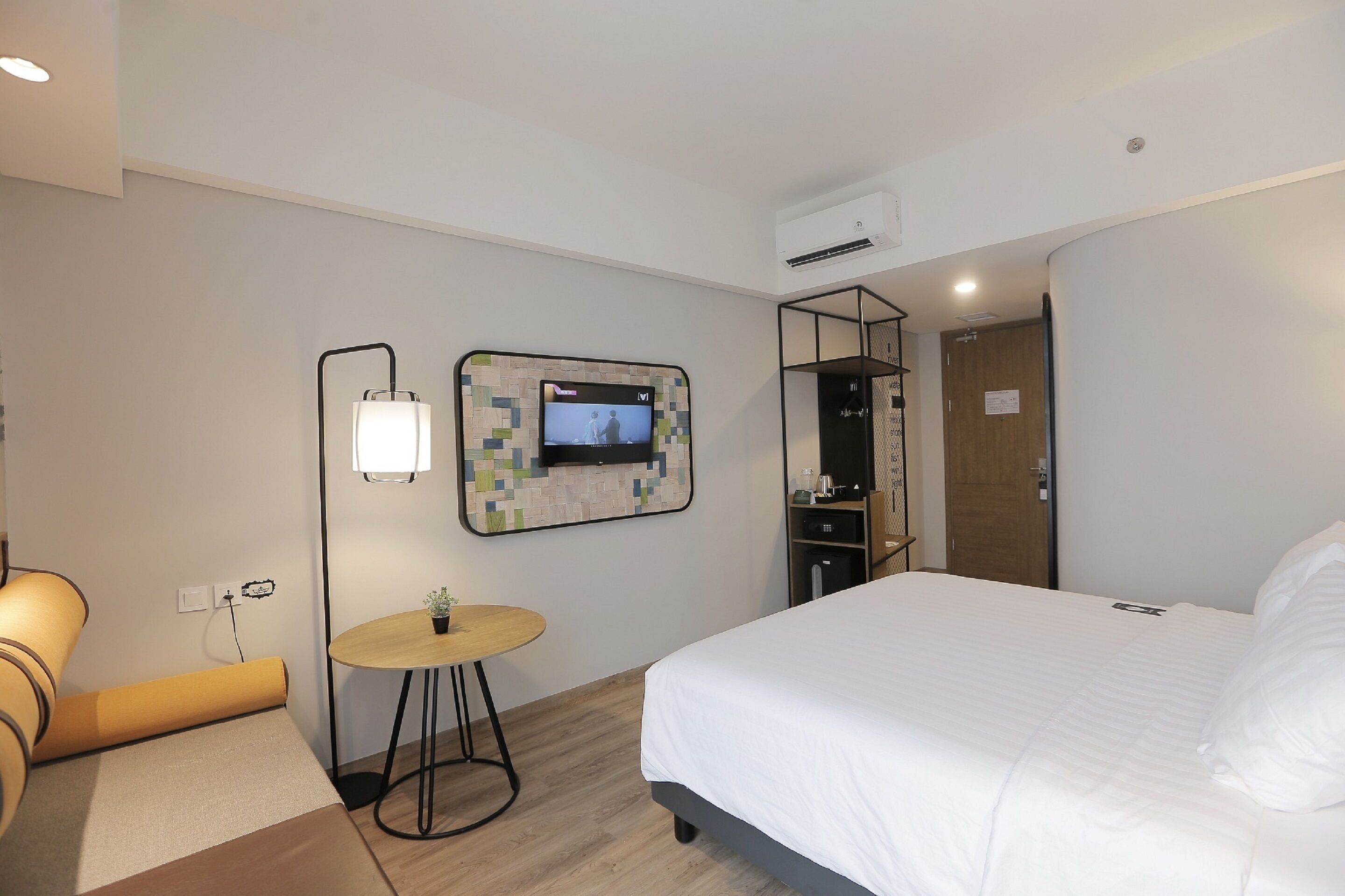 Quarto de hotel com uma cama, uma mesinha redonda com abajur, uma TV fixada na parede e um guarda-roupa.