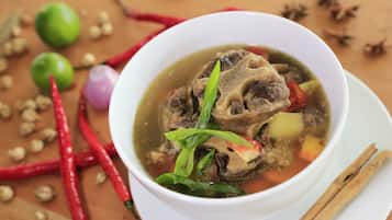 A bowl of soup with meat and vegetables, surrounded by red chili peppers and a lime.