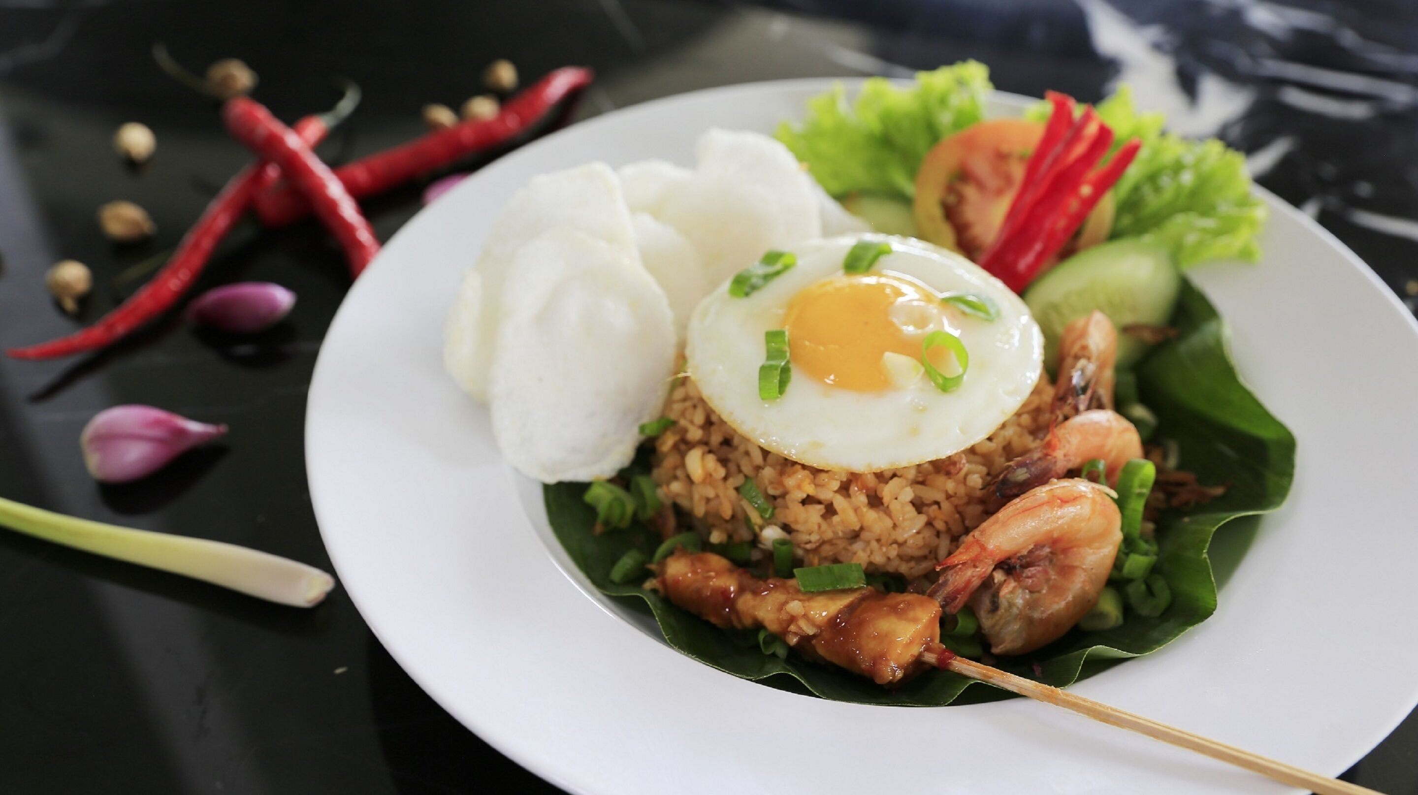A plate of fried rice topped with a fried egg, shrimp, and a white dumpling, served on a bed of lettuce with a side of sliced cucumber and tomato.