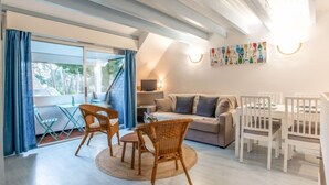 Un salon moderne comprenant un canapé, deux fauteuils, une table à manger et un téléviseur. Il y a une grande fenêtre avec des rideaux, un luminaire fixé au mur et une œuvre d’art aux couleurs vives.