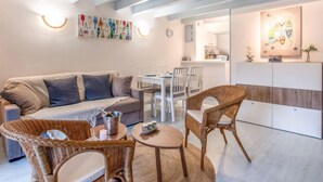 Un salon chaleureux avec un canapé, deux fauteuils en osier, une petite table ronde et, au fond, un coin cuisine.