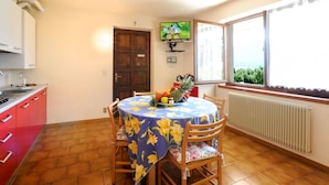 A dining area with a round table covered in a floral tablecloth, surrounded by wooden chairs. A fruit bowl is on the table, and a television is mounted on the wall above it.