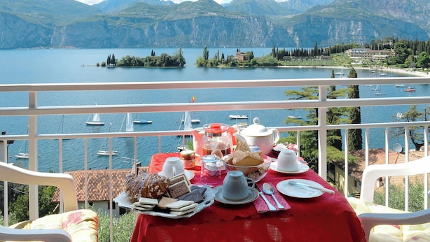 A table set with breakfast items on a balcony overlooking a lake with boats and mountains in the background.