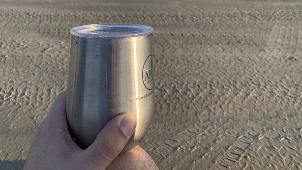 Una mano sosteniendo una lata en una playa arenosa, con el océano al fondo.