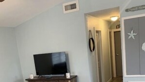 Una sala de estar con una televisión de pantalla plana sobre un soporte de madera, un pasillo que lleva a una puerta y un banco en la esquina.