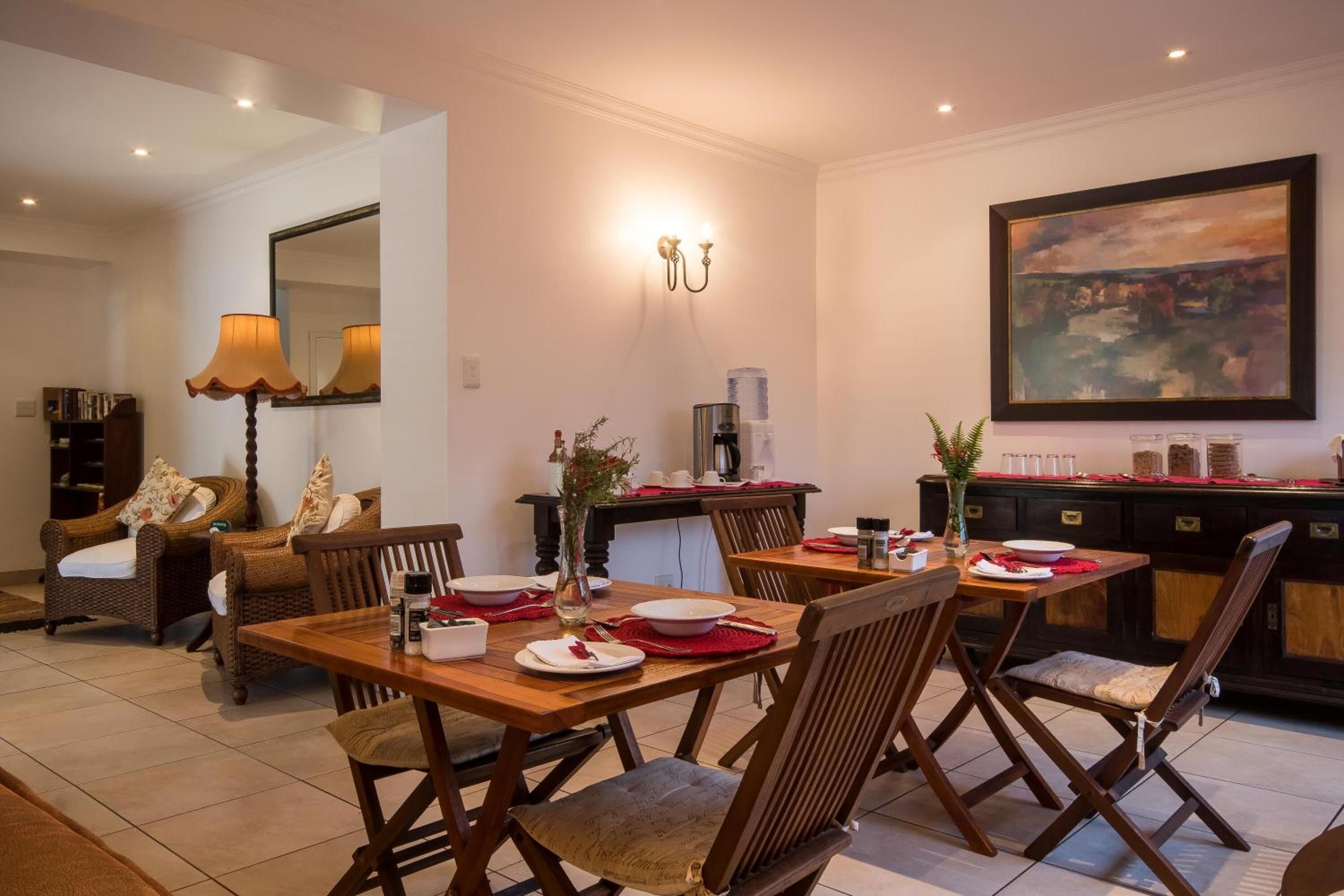 A dining area with a wooden table set for four, surrounded by wicker chairs. A mirror and wall-mounted sconces provide lighting.