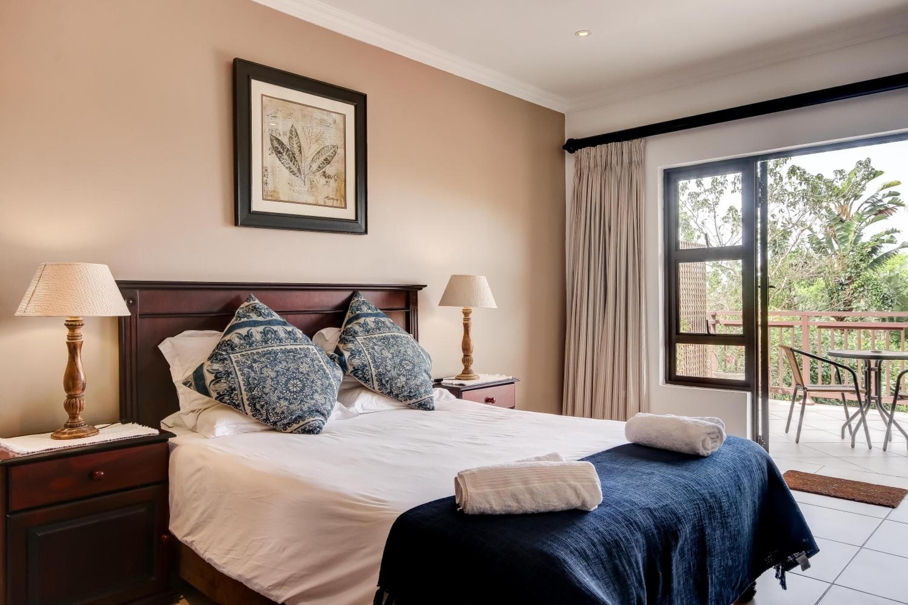 A hotel room with a large bed, two bedside lamps, a framed artwork above the bed, and a view of the outdoors through a sliding glass door.