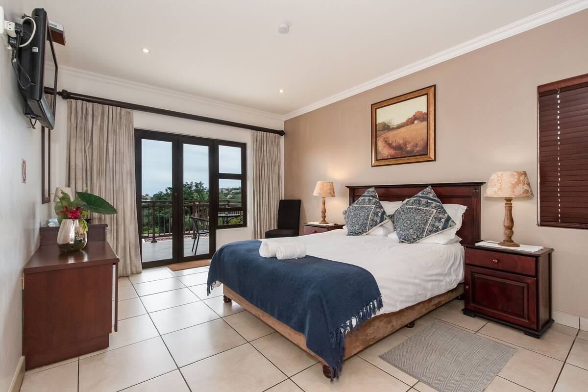 A bedroom with a large bed, two bedside tables, a TV mounted on the wall, a painting above the bed, and a view of the outdoors through a sliding door.